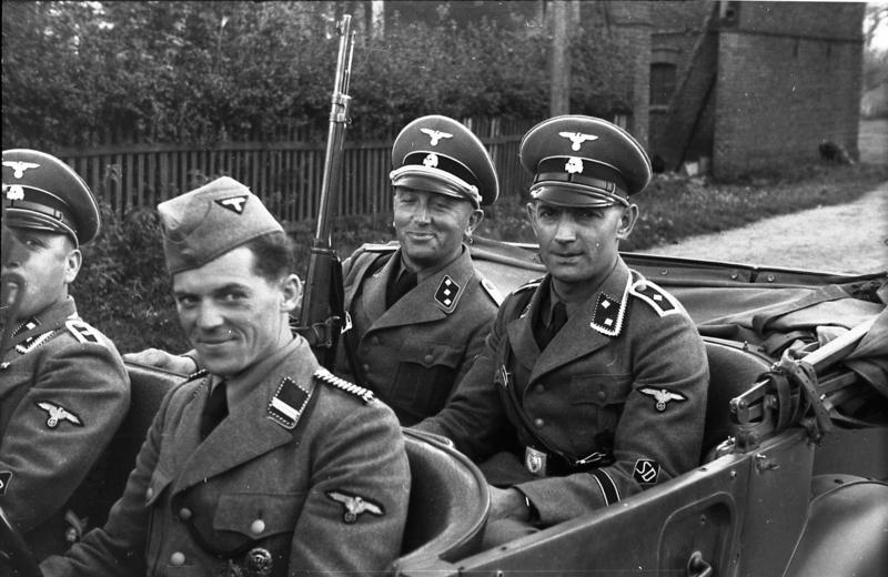1939 szeptember, Lengyelrszág, Ustronice / Opatowice közelében; a lengyel és a náci Németország biztonsági rendőrsége felügyelete alatt álló letartóztatott zsidók úti ellenőrzése. Négy rendőr ül egy kabrioletben: balról jobbra, egy Oberscharführer, egy Rottenführer egy Untersturmführer és egy Oberscharführer. Forrás: Deutsches Bundesarchiv. 